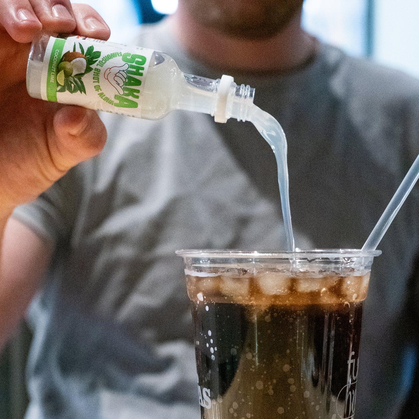A person in a gray shirt pours a small bottle of Shaka Beverage Company’s Coconut Lime into a plastic cup holding a dark iced drink with a straw, adding a refreshing tropical twist.