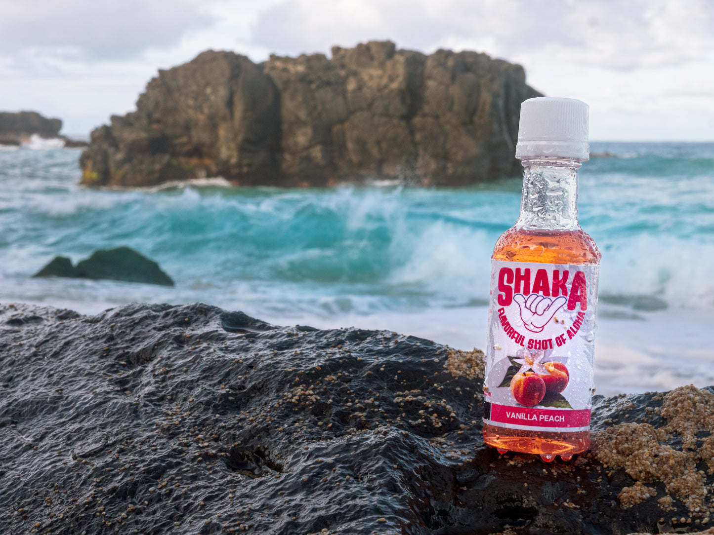 A bottle of Shaka Beverage Company Vanilla Peach drink mix sits on a black rock at the beach, with turquoise waves and a large rocky formation in the background.