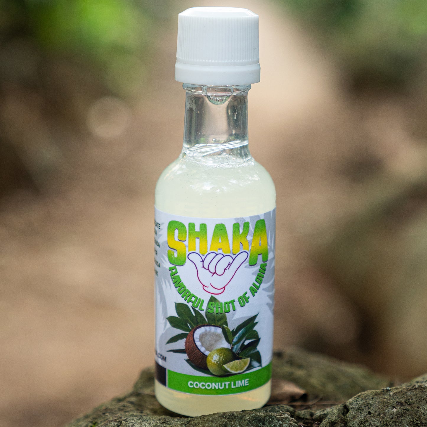 A small clear bottle labeled Shaka Beverage Company Coconut Lime, with coconut and lime images, sits on a rock outdoors, evoking a fresh tropical vibe against a blurred natural background.